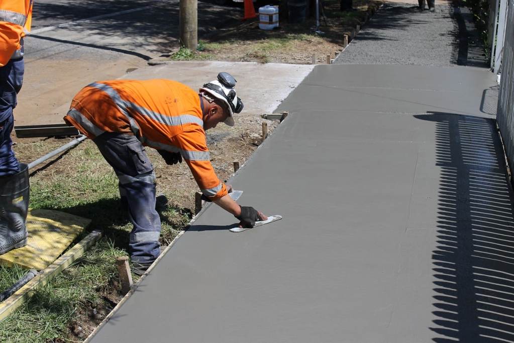 Worker wearing orange hi-vis jacket and white hard hat, beding over freshly laid concrete with a small hand-held tool to level the surface